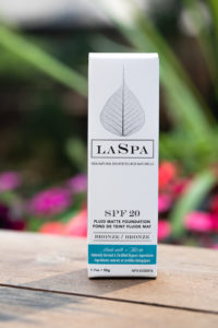 Product box of LASPA's SPF 20 tinted mineral sunscreen standing on a wood table with pink flowers in background