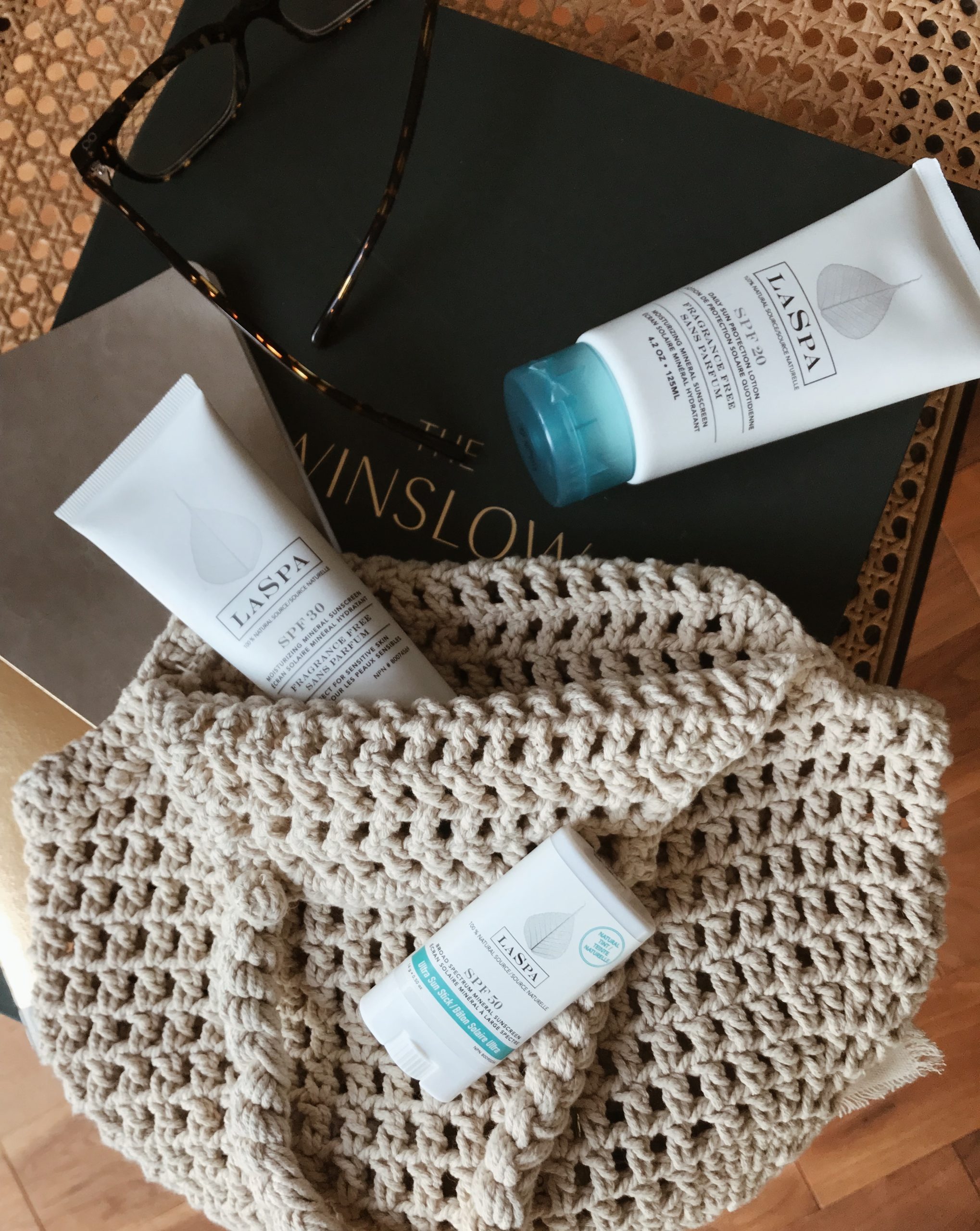 Three white bottles of LASPA Mineral SPF Sunscreen products on a table with a beige bag, a black book and sunglasses.